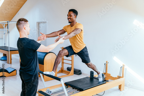 Fitness instructor corrects and controls the Pilates exercise that his african american male student is doing on Reformer bed in health center, gym with modern interior.