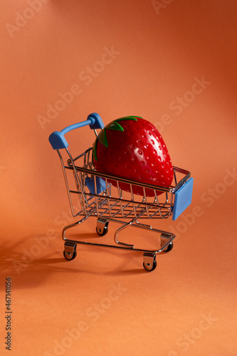 Large plastic strawberry in a small supermarket trolley. Fruit delivery. Fruit cart on a beige background. 