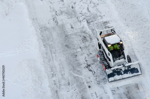 Skid steer loader removes snow from the city streets. Top view of the road with cars and snow blower. Seasonal work in winter snowy city. Equipment and city worker.