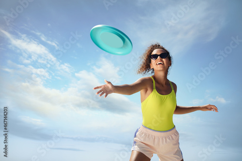 Happy African American woman throwing flying disk against blue sky on sunny day