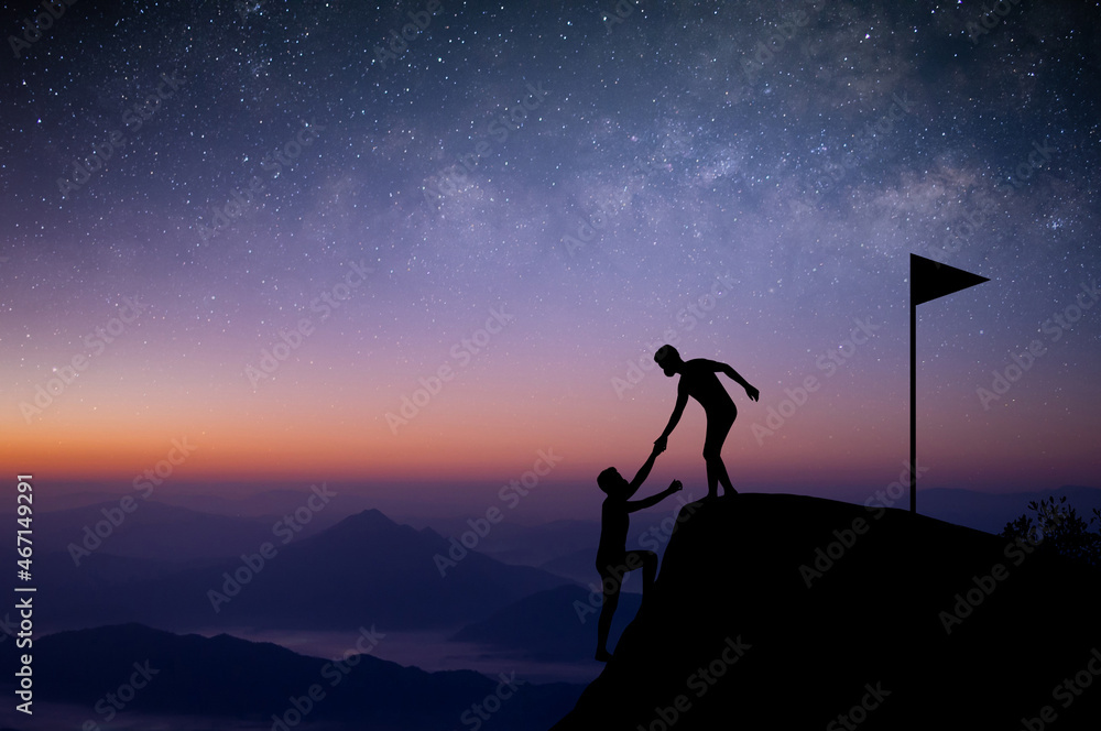 Two tourists are climbing with a friend pulling their hands before reaching the summit with the beautiful night sky, stars, the Milky Way in the background.