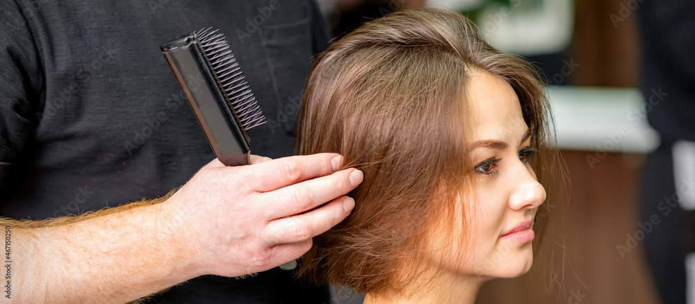 Fototapeta premium Male hairdresser works on the hairstyle of the young caucasian brunette woman at a hair salon