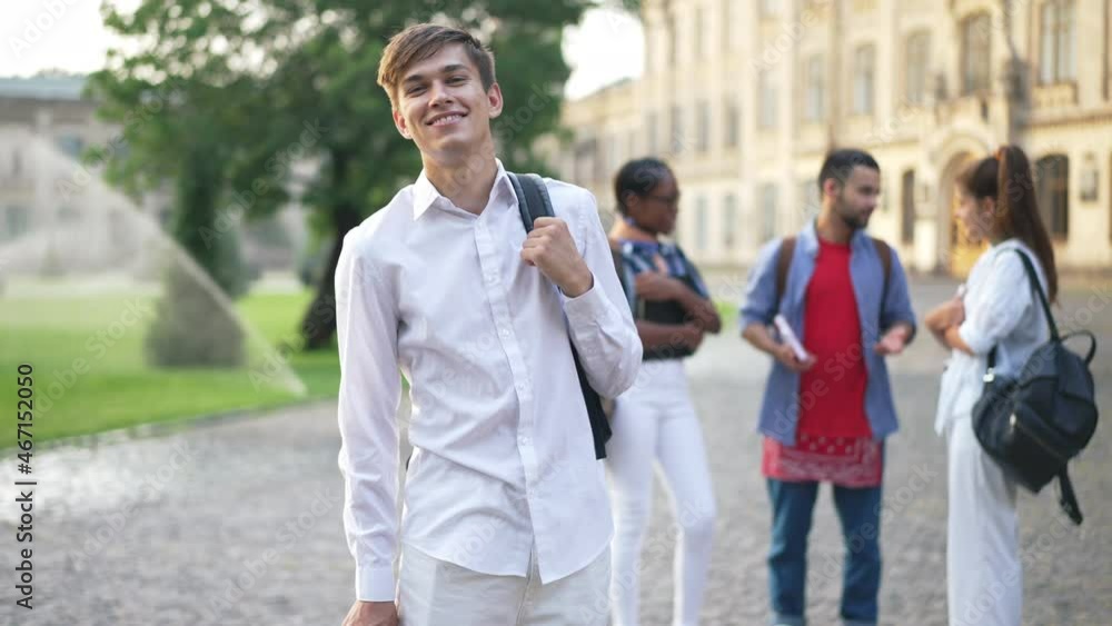 Portrait of positive confident young Caucasian man hanging backpack on shoulder smiling looking at camera. Blurred multiracial groupmates standing at background talking in slow motion. Education Portrait of positive confident young Caucasian man hanging backpack on shoulder smiling looking at camera. Blurred multiracial groupmates standing at background talking in slow motion. Education