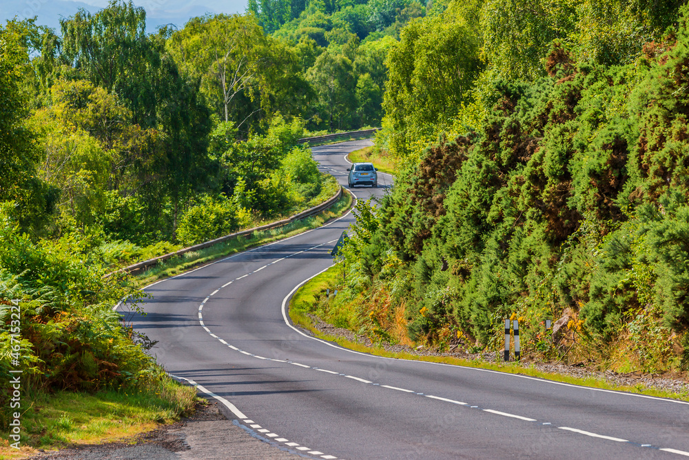 A car drives on a road. A narrow, winding road in Scotland along Loch Ness. Trees and bushes next to the road in summer in sunshine. Traffic signs and guard rails