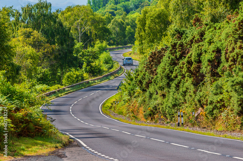 Obraz na płótnie A car drives on a road. A narrow, winding road in Scotland along Loch Ness. Trees and bushes next to the road in summer in sunshine. Traffic signs and guard rails w salonie