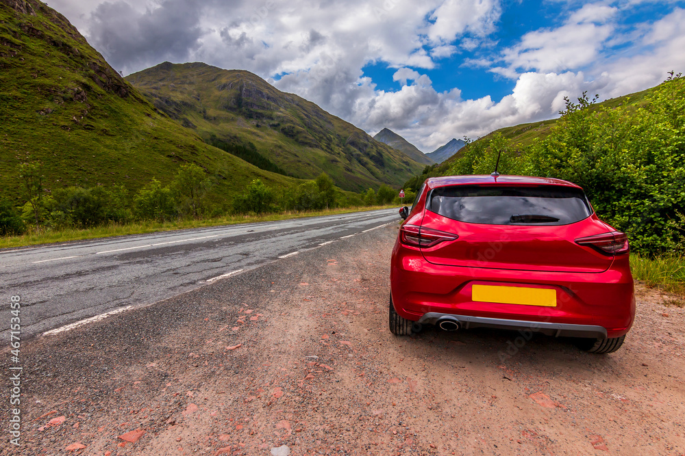 Naklejka premium Mountains in the Scottish Highlands. Road through the countryside with a red car in the foreground. Clouds in the sky in the daytime with sunshine. Green trees and bushes