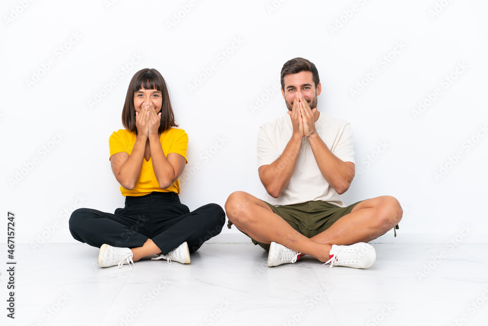 Young couple sitting on the floor isolated on white background smiling a lot while covering mouth Young couple sitting on the floor isolated on white background smiling a lot while covering mouth
