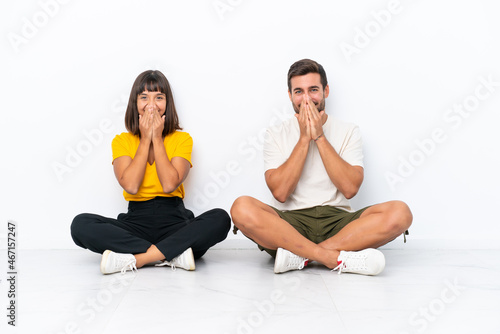 Young couple sitting on the floor isolated on white background smiling a lot while covering mouth