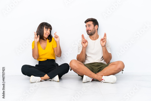 Young couple sitting on the floor isolated on white background with fingers crossing and wishing the best
