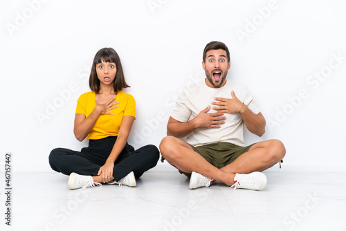 Young couple sitting on the floor isolated on white background surprised and shocked while looking right