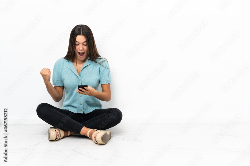 Teenager girl sitting on the floor surprised and sending a message