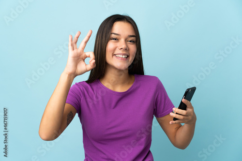 Teenager girl isolated on pink background using mobile phone and doing OK sign