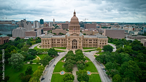 Aerial view of Texas State Capitol building in Austin, surrounded by lush greenery & city skyline under cloudy skies. Iconic landmark!