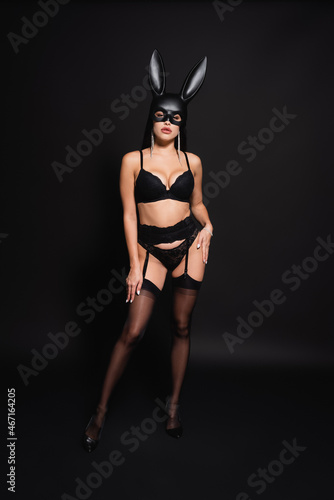 full length view of sexy woman in lingerie, heels and bunny mask looking at camera while posing on black