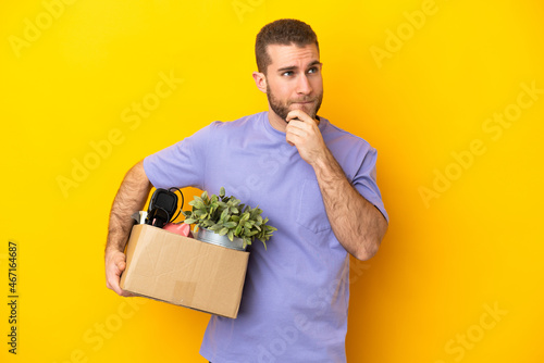 Young caucasian making a move while picking up a box full of things isolated on yellow background having doubts and thinking