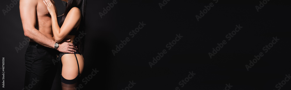 Fototapeta premium partial view of shirtless man hugging woman in lingerie and stockings isolated on black, banner