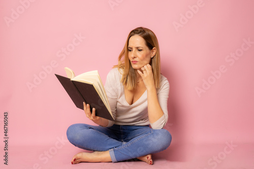 Young woman sitting on floor reading a book isolated over pink background.