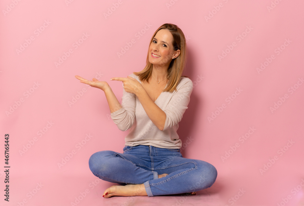 Smiling woman sitting on floor cross-legged pointing finger side isolated over pink background. Smiling woman sitting on floor cross-legged pointing finger side isolated over pink background.