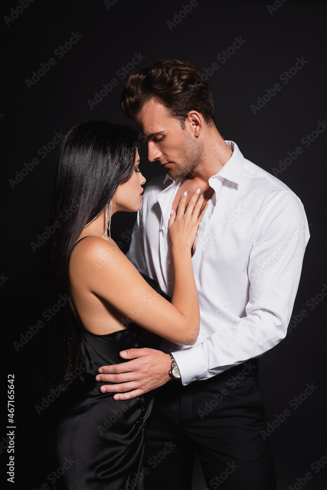 young man in unbuttoned shirt hugging elegant woman in silk dress isolated on black