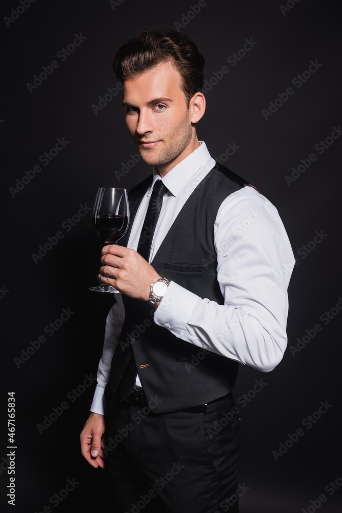positive and confident man looking at camera while holding glass of red wine isolated on black positive and confident man looking at camera while holding glass of red wine isolated on black