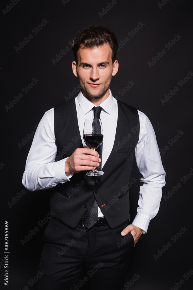 front view of smiling and elegant man holding glass of red wine while standing with hand in pocket isolated on black front view of smiling and elegant man holding glass of red wine while standing with hand in pocket isolated on black