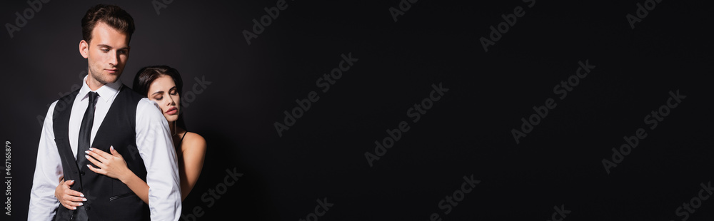 sensual woman with closed eyes hugging stylish man isolated on black, banner