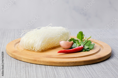 Dry funchose and ingredients for preparing spicy Asian dish. Chinese glass noodles, garlic, pods of red hot pepper and cilantro on a wooden cutting board. Cooking vegetarian meals. photo