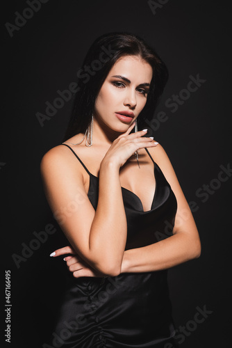 elegant brunette woman in dress holding hand near face while looking at camera isolated on black