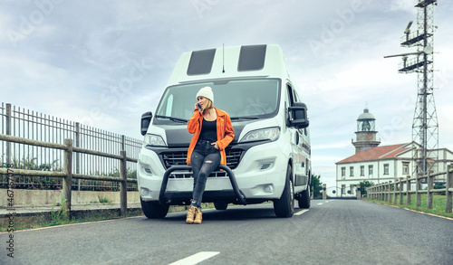 Young woman leaning on camper van on the road calling by mobile for a breakdown with lighthouse in background