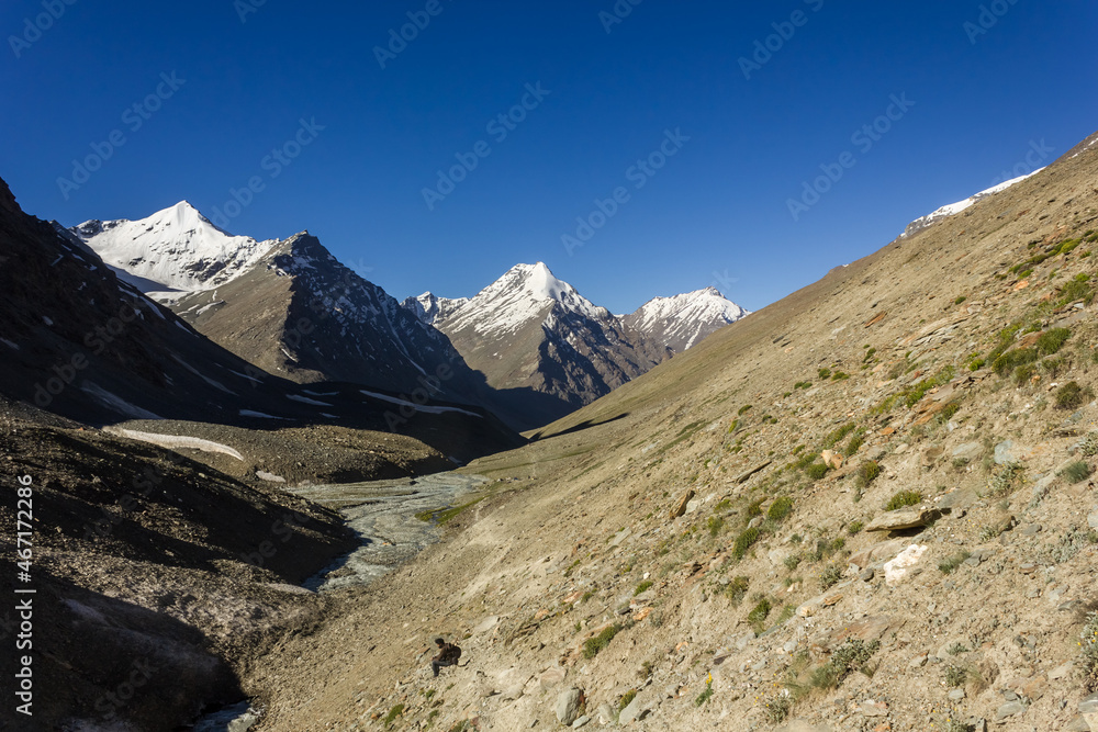 A view of snow capped Himalayan mountains under a blue sky from a trekking trail on the steep slope of a hill in Zanskar.