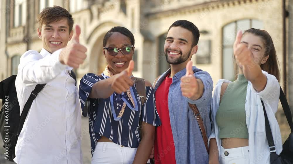 Medium shot four happy students looking at camera smiling showing thumbs up simultaneously. Portrait of confident smart multiethnic men and women posing on university yard at campus outdoors Medium shot four happy students looking at camera smiling showing thumbs up simultaneously. Portrait of confident smart multiethnic men and women posing on university yard at campus outdoors