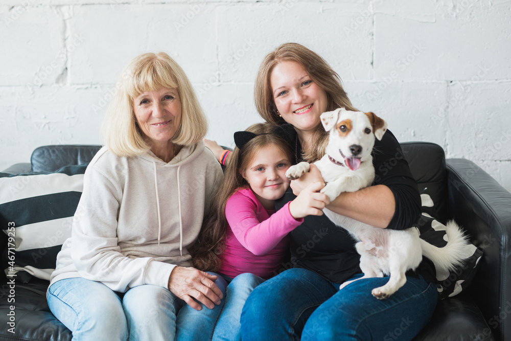 Happy granddaughter and grandmother and daughter sitting on sofa with jack russell terrier dog. Grandma hugs grandchild at home. Relationship, family and three generation concept. Happy granddaughter and grandmother and daughter sitting on sofa with jack russell terrier dog. Grandma hugs grandchild at home. Relationship, family and three generation concept.
