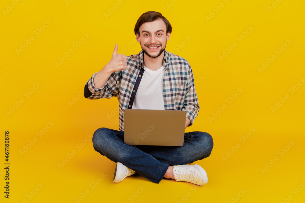 Full size photo of a happy young man holding laptop computer show thumb-up while sitting on a floor isolated over yellow color background
