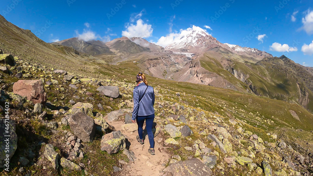 A woman walking on a narrow path towards Mount Kazbeg in Caucasus, Georgia. There slopes are overgrown with green grass. Massive Gergeti Glacier under the sharp peak. Cloudless sky. Exploring.