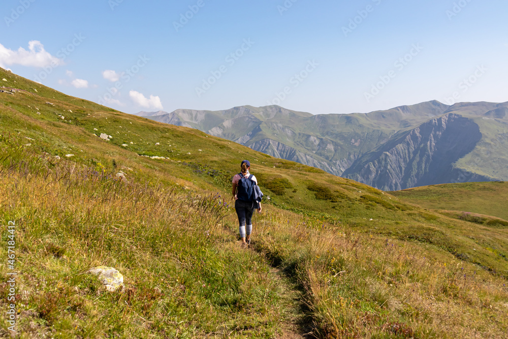 A woman hiking along a narrow pathway in high Caucasus mountains in Georgia. There are high glaciers in the back. Thick clouds above the sharp peaks. Lush pastures on the sides. Barren peaks.
