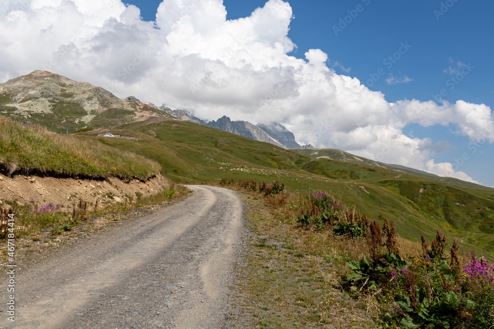 A gravelled road in high Caucasus mountains in Georgia. The road leads to a ski resort on a steep slope. Thick clouds in the back. Lush pastures on the sides. Barren peaks.