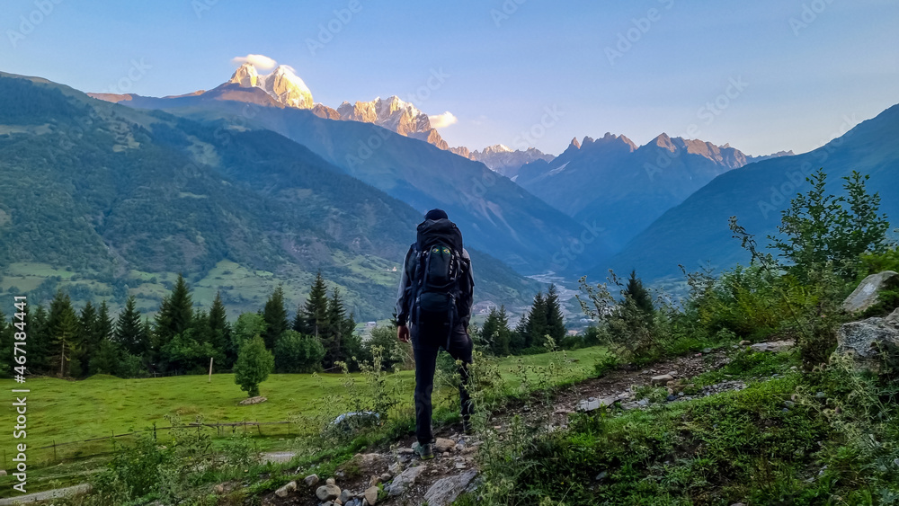 A man with big hiking backpack standing on a lush pasture with a view on the first sunbeams reaching the peaks of Ushba in Caucasus, Georgia. Cloudless sky above the high and snow-capped mountains.