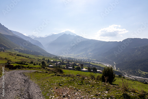 A gravel road leading to Zhabeshi, a mountain village in Georgia. A few lookout towers towering above the village. High Caucasus mountain chains. There is a river flowing through the valley. photo