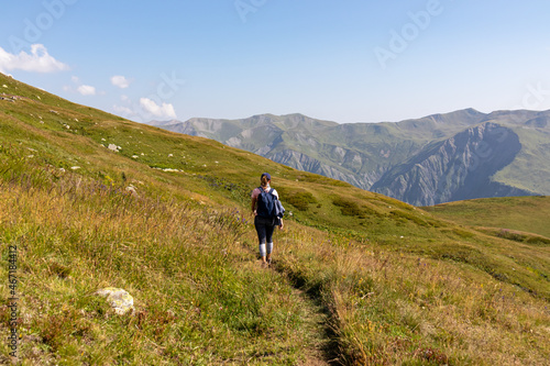 A woman hiking along a narrow pathway in high Caucasus mountains in Georgia. There are high glaciers in the back. Thick clouds above the sharp peaks. Lush pastures on the sides. Barren peaks. © Chris
