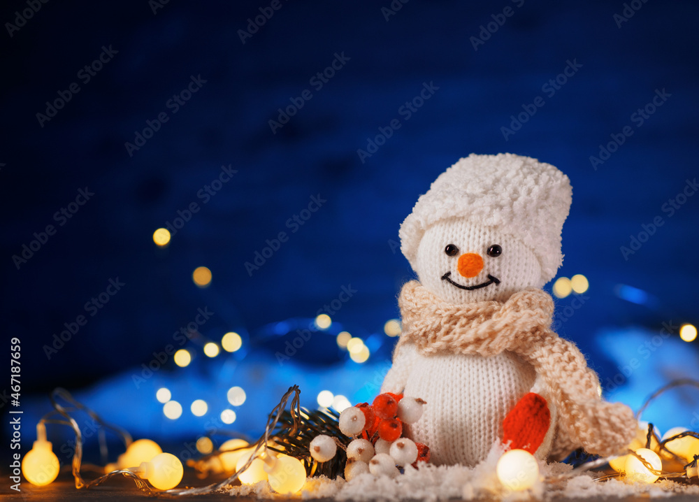 Obraz premium Funny knitted toy snowman. Christmas decor, fake snow decoration , bokeh garlands, blue background copy space