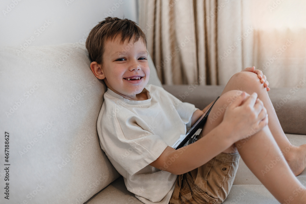 Little boy using digital tablet on bed during the day. Growing up in a technology-based world. Shot of a young boy using his digital tablet while lying on the sofa, looking at camera and smiling. Little boy using digital tablet on bed during the day. Growing up in a technology-based world. Shot of a young boy using his digital tablet while lying on the sofa, looking at camera and smiling.