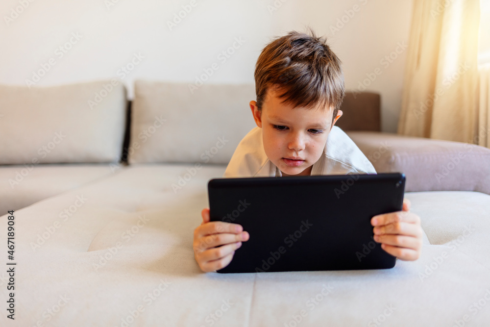 Modern kid and education technology. Adorable little Caucasian boy sitting on sofa and playing game on digital tablet. Shot of a young boy using a digital tablet in bed during the day at home. Modern kid and education technology. Adorable little Caucasian boy sitting on sofa and playing game on digital tablet. Shot of a young boy using a digital tablet in bed during the day at home.