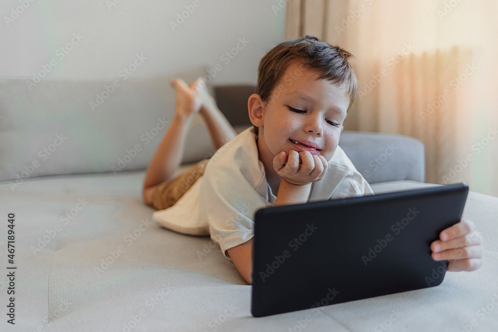 Little cute boy in a beige T-shirt playing games on a tablet and watching cartoons. Toddler with digital tablet at home. Technology concept. Child with a gadget. Copy space. Little cute boy in a beige T-shirt playing games on a tablet and watching cartoons. Toddler with digital tablet at home. Technology concept. Child with a gadget. Copy space.
