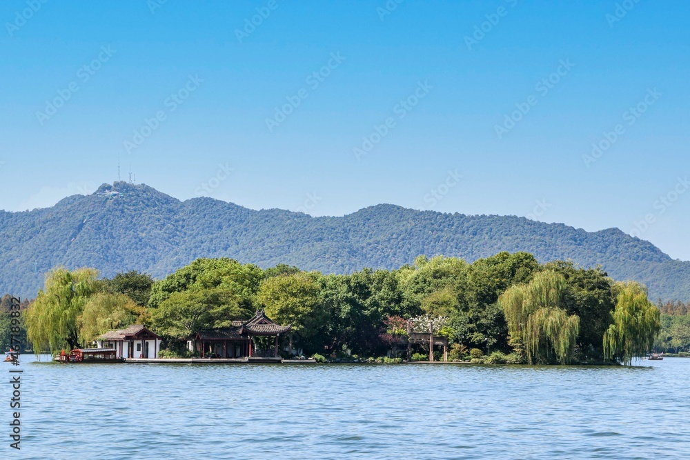 A small green island on the West Lake in Hangzhou, China, filled with green trees and surrounded by hills, mountains and a blue sky A small green island on the West Lake in Hangzhou, China, filled with green trees and surrounded by hills, mountains and a blue sky