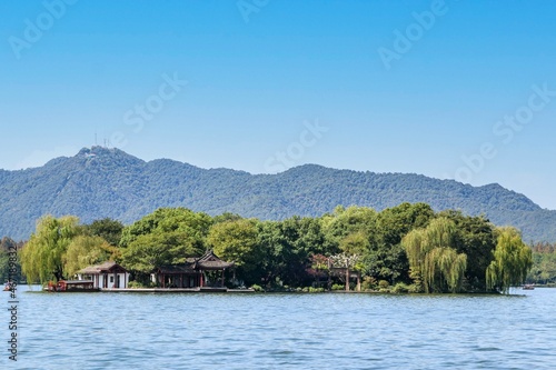 A small green island on the West Lake in Hangzhou, China, filled with green trees and surrounded by hills, mountains and a blue sky