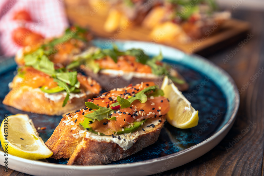 delicious bruschetta with salmon, avocado and arugula with lemon wedges on a large blue plate on a woody background.