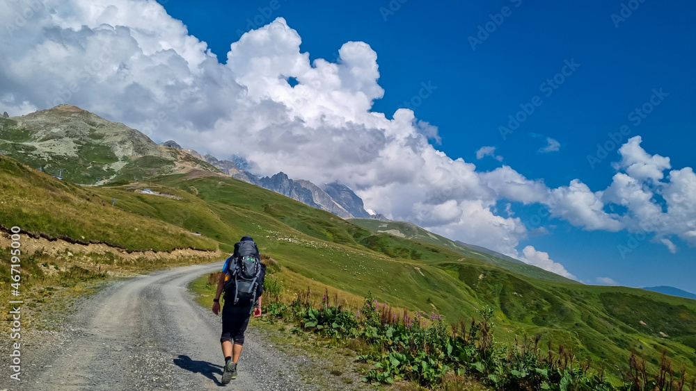 Fototapeta premium A man hiking along a gravelled road in high Caucasus mountains in Georgia. The road leads to a ski resort on a steep slope. Thick clouds in the back. Lush pastures on the sides. Barren peaks.