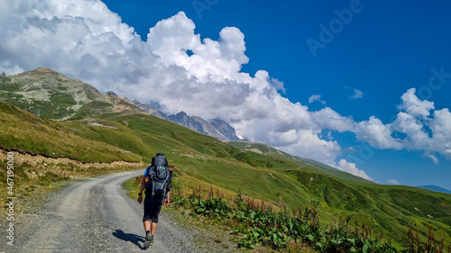 A man hiking along a gravelled road in high Caucasus mountains in Georgia. The road leads to a ski resort on a steep slope. Thick clouds in the back. Lush pastures on the sides. Barren peaks. photo