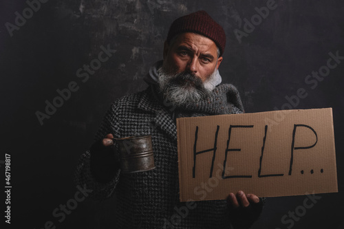 Portrait of poor homeless man with poster and a pot for alms on a dark background