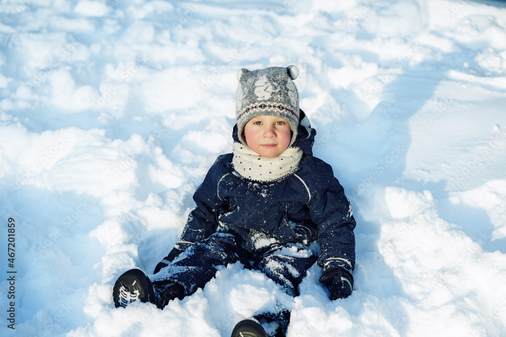 cute caucasian boy in warm winther jacket, scandinavian hat and scarf sitting in snow in city park on winter day cute caucasian boy in warm winther jacket, scandinavian hat and scarf sitting in snow in city park on winter day